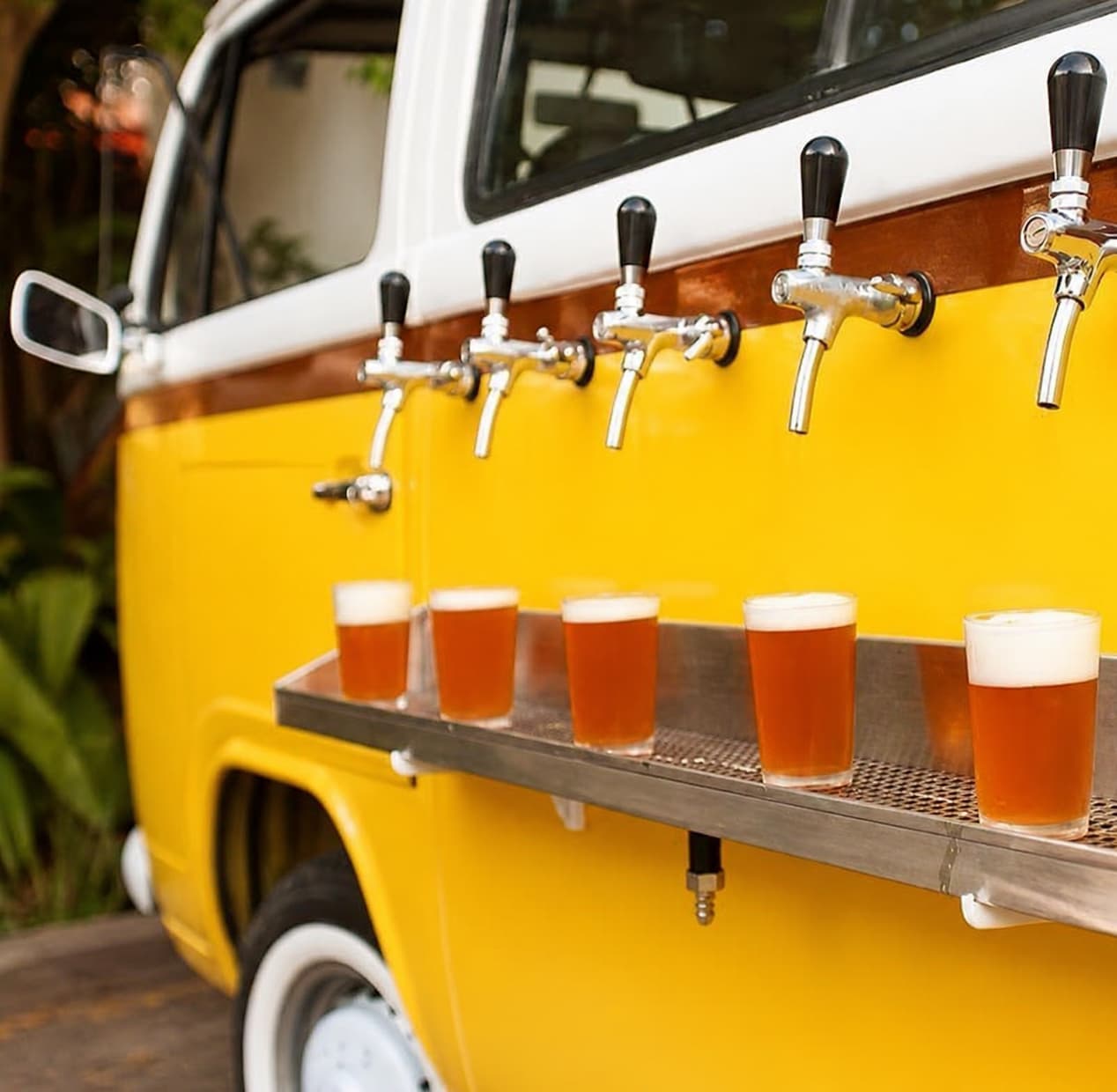 Yellow VW T2 with six beer taps and row of pint glasses on steel shelf in tropical garden