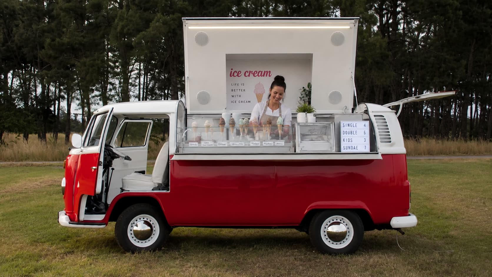 Red and white VW T2 ice cream truck with smiling woman serving cones from glass display case